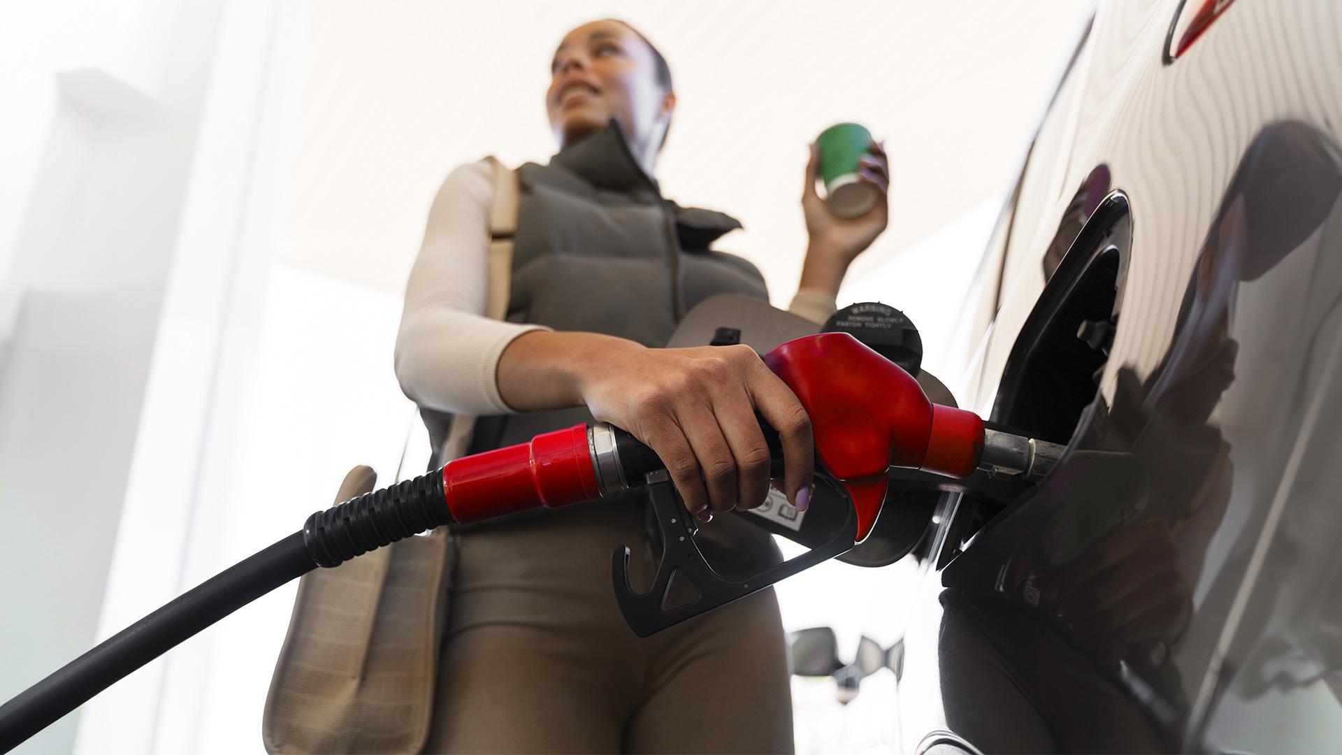 A woman fills up her car with gas with her right hand and holds a cup in the other.