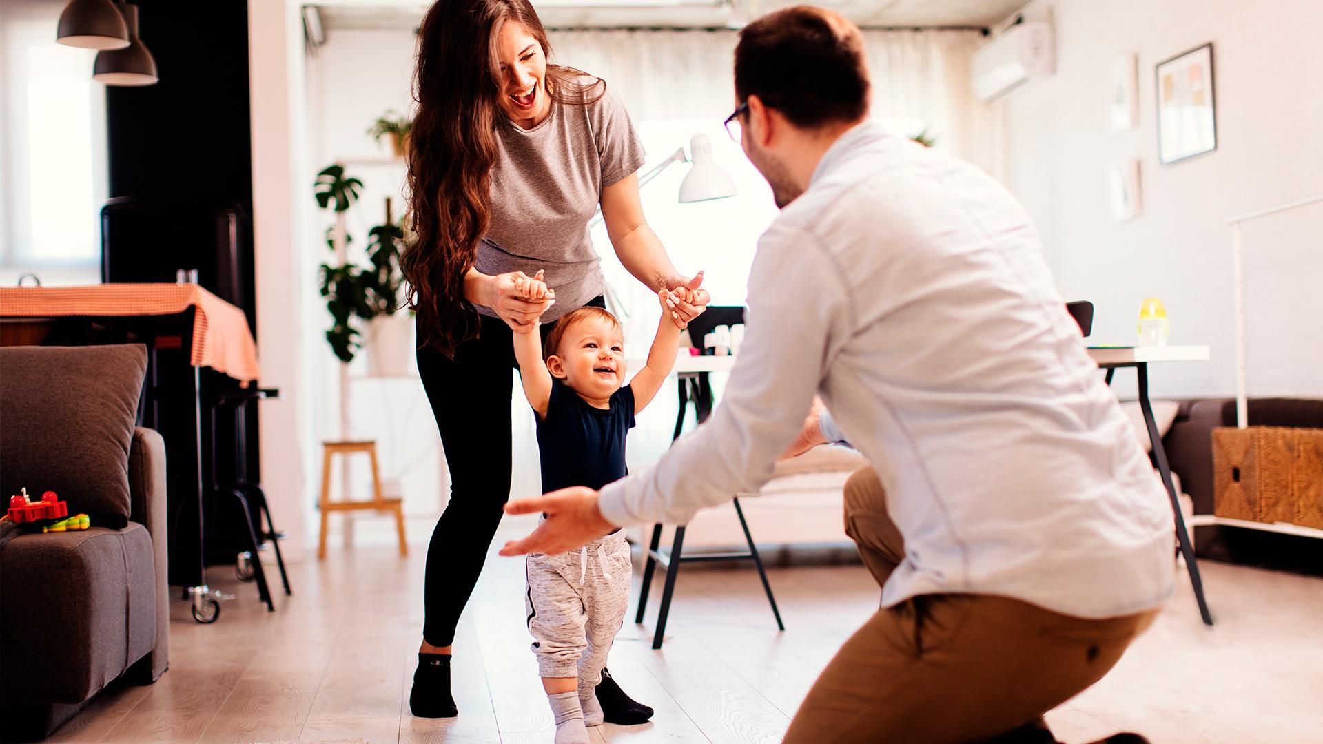 A woman holds her son while his father encourages him to walk.