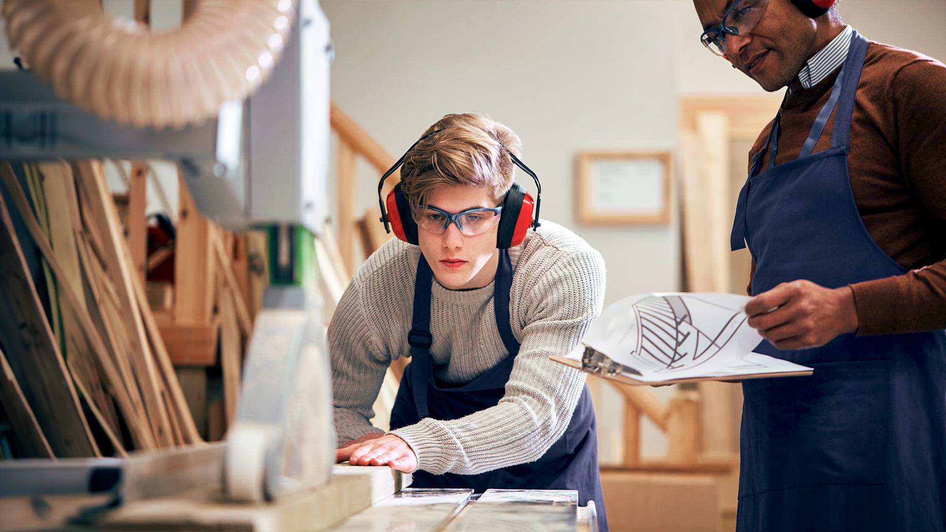A young student cuts wood in a woodworking class while his teacher observes.